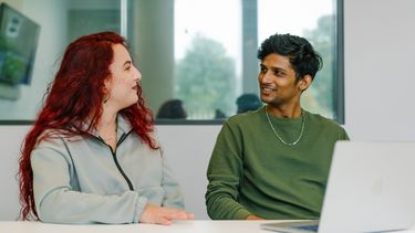 Two students siting at a table in front of a laptop talking