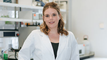 Rachel stands in her lab, hands in her lab coat pocket smiling at the camera